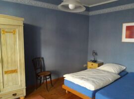 Ein Schlafzimmer mit zwei zusammengestellten Einzelbetten auf der rechten Seite. Darüber ein Bild an der Wand und ein kleiner gelber Nachttisch mit Lampe links daneben. Links ein heller Kleiderschrank und ein Holzstuhl.