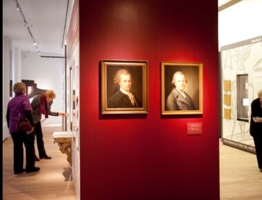 Innenansicht des Lessing-Museums mit zwei Gemälden im Mittelpunkt, auf denen der Aufklärer und Schriftsteller Lessing zu sehen ist. Drei Besucherinnen sehen sich die weiteren Ausstellungsstücke rechts und links im Raum an.