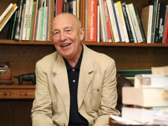 Foto-Portrait des Künstlers Georg Baselitz als Text-Ergänzung. Der ältere Herr sitzt vor einem Bücherregal und lächelt in die Kamera. Er trägt ein schwarzes Hemd und ein sandfarbenes Sakko darüber. Im Vordergrund sind unscharf Bücher zu erkennen.