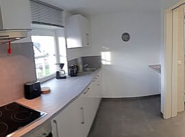Helle moderne Küchenzeile unter einem Fenster. Links Herd mit Ofen und Abzugshaube. Nach rechts Toaster, Kaffeemaschine, Wasserkocher, Spüle und Schränke. Rechts Eingangstür.