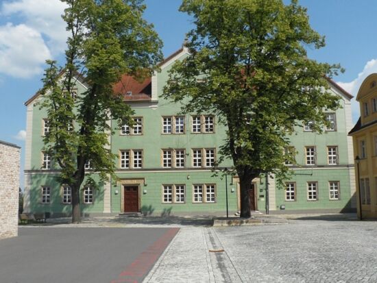 Außenansicht der "Grünen Schule" auf dem Schulplatz bei Sonnenschein. Ein mittelgroßes grünes Gebäude mit zwei Eingangspforten. Über beiden prangt eine goldene Inschrift aus der Ferne.