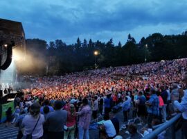 Ein riesiges Publikum vor der Hutberg-Freilichtbühne in Kamenz während eines Konzerts. Im Hintergrund umranden Bäume des Hutbergparks das Gelände. Am Himmel ziehen Wolken zur Abenddämmerung vorüber.