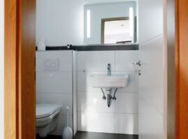 Badansicht mit Toilette links und Waschbecken mit beleuchtetem Spiegel rechts.