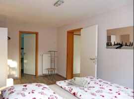 Ein helles Schlafzimmer mit einem großen Doppelbett und Blümchenbettwäsche. Links ein Kleiderschrank und Zugang zum Bad. Rechts ein Stuhl / Kleiderbuttler und Eingangstür. An der Wand ein Spiegel mit Toskana-Silhouette.