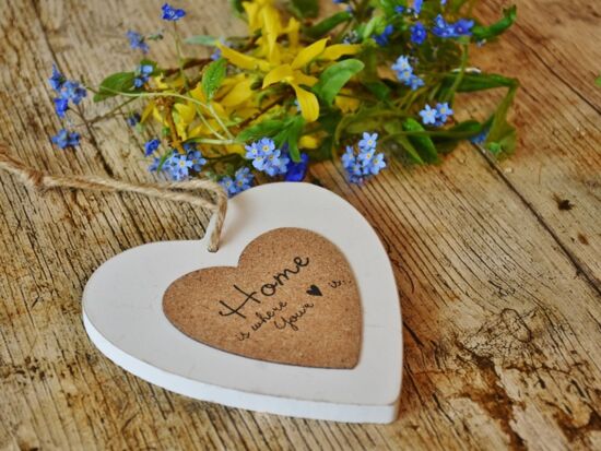 Ein Holz- / Korkherz mit der Aufschrift "Home is where your heart is" übersetzt "Zuhause ist, wo das Herz ist". Es liegt auf einem Holztisch mit Wildblumen. Symbolbild für Wohnen und Heim.Holztisch