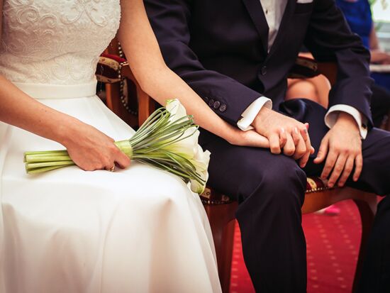 Symbolbild für eine Trauung. Ein Traupaar sitzt gemeinsam auf Stühlen, Ausschnitt mit Beinen und Oberkörper des Paares. Braut im weißen Kleid links hält einen Strauß Blumen. Bräutigam in Anzug hält ihre Hand.