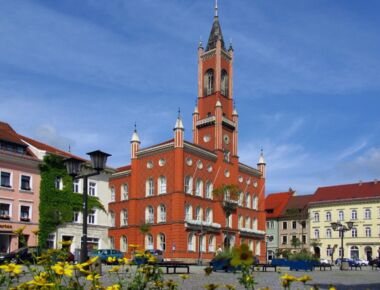 Marktplatz in Kamenz mit Rathaus im Zentrum und Wohn- und Geschäftshäusern drumherum. Aufnahme bei Sonnenschein mit Blumenschmuck im Vordergrund. Beispiel für Sehenswürdigkeiten in Kamenz.
