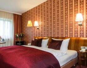 Einblick in ein Doppelzimmer des Hotels Goldner Hirsch in Kamenz. In der Mitte ein großes, gemütlich wirkendes Bett. Rechts und links stehen Nachttische mit Blumen und Kärtchen. Das Zimmer ist in edlen Rot- und Goldtönen gehalten. Übernachtungsbeispiel.