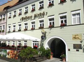 Vorderansicht des Eingangsbereiches des AKZENT Hotel Goldner Hirsch. Rechts das Goldrelief eines Hirschen. Links eine Terrasse mit Stühlen, Tischen und Schirmen.