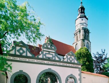 Eingangsportal der Hauptkirche St. Marien in Kamenz. Im Vordergrund ein Tor mit drei Rundbögen und der Aufschrift "Der Herr behüte deinen Ausgang und Eingang von nun an bis in Ewigkeit".